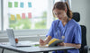 A female nurse in scrubs sits at a desk with a notebook, preparing for the NCLEX exam with practice questions.